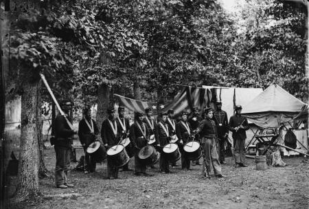 The drum corps poses with their drums, in camp beside a draped American flag and tents.