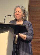 Valerie stands at a podium with a microphone and smiles at her audience. She is wearing a long, flowing purple-grey cardigan over a white shirt and black pants.