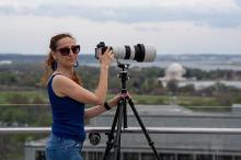 Yuli stands with her camera on a tripod, with DC monuments in the background.