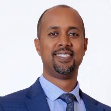 Headshot of Noah Abraham, Director of the Center for Operations at DCHS. He is a smiling man with a short beard and mustache, wearing a blue suit jacket and a white dress shirt, against a light gray background.