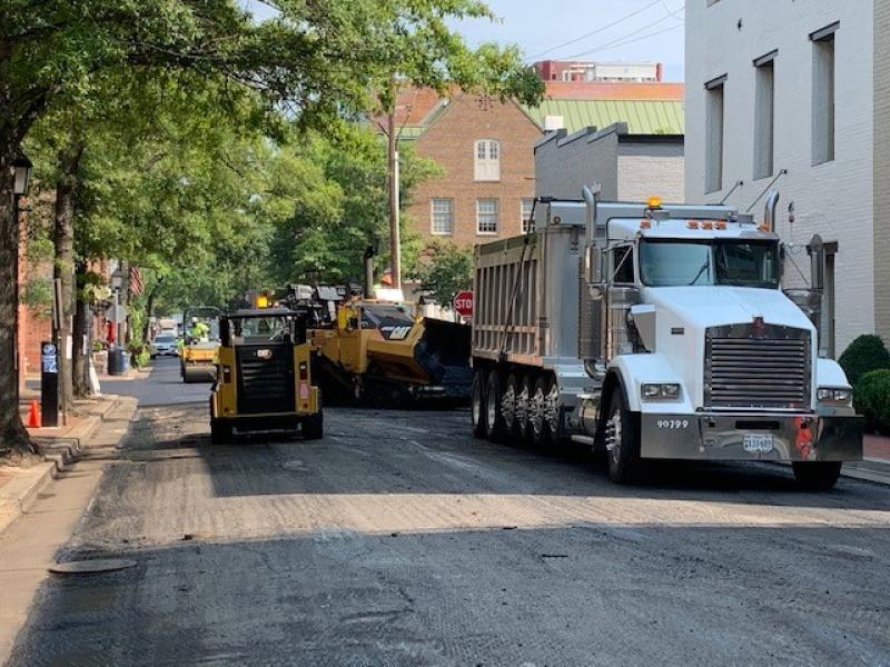 A white truck carrying asphalt and a bobcat vehicle repave an Alexandria street
