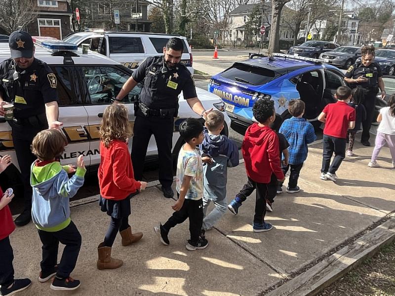 A line of several elementary school children walking past three uniformed deputies and giving high fives. Children are shown from behind and marked Sheriff's Office vehicles are in the background.