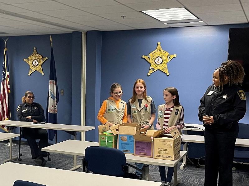 Three Girl Scout Cadettes delivering four cartons of cookies to two deputies in a roll call room